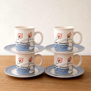 May include: Four white ceramic mugs with a pink floral design and matching saucers. The mugs have a curved handle and are stacked in pairs. The saucers are light blue with a white centre and a blue rim. The mugs and saucers are arranged on a wooden surface.