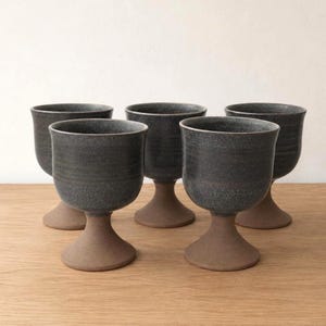 Pode incluir: Um conjunto de cinco cálices de cerâmica com um esmalte cinzento-carvão salpicado na taça superior e uma base de argila castanha mate não vidrada. Cada cálice tem uma forma arredondada, um pé curto e uma base circular larga e estável.