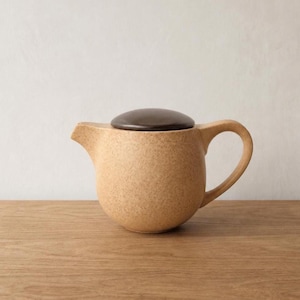 May include: A beige ceramic teapot with a dark brown lid. The teapot has a rounded body, a curved handle, and a spout. The surface has a speckled texture. The teapot is on a wooden surface.