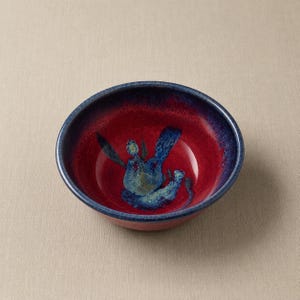 May include: A round ceramic bowl featuring a deep red interior with a dark blue rim and a central abstract blue and green glaze pattern. The exterior of the bowl is a solid dark blue, creating a contrast with the vibrant red and blue interior finish.