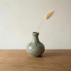 May include: A small, sage green ceramic vase with a rounded body and narrow neck. The vase has subtle, dark markings and holds a single, dried, fluffy beige flower. The vase sits on a wooden surface.
