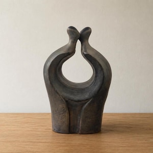 May include: A dark grey ceramic sculpture of two embracing figures, with a large open space in the centre. The sculpture has a smooth, matte finish and sits on a wooden surface.