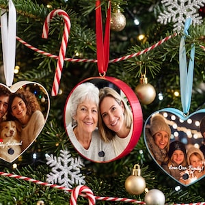 Op de afbeelding: Drie gepersonaliseerde foto-ornamenten hangen in een kerstboom. Een hartvormig ornament met een wit lint, een rond rood ornament met een rood lint en een lichtblauw hartvormig ornament met een lichtblauw lint, elk met een familiefoto.