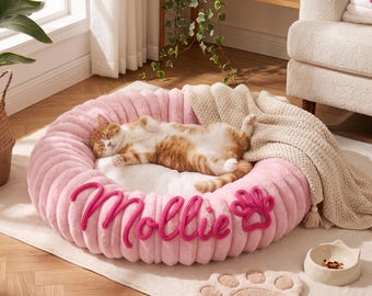 May include: A round, pink, ribbed pet bed with a white plush center, featuring the name Mollie and a paw print embroidered in dark pink. A ginger and white cat sleeps inside the bed, which is draped with a beige knit throw blanket.