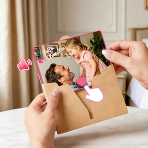 Peut inclure: Une carte de vœux rectangulaire présentant une photographie d'un homme tenant un jeune enfant. La carte a une bordure rose et le texte blanc "Papa I love you!" imprimé sur l'image. Elle est partiellement insérée dans une enveloppe en papier brun clair.