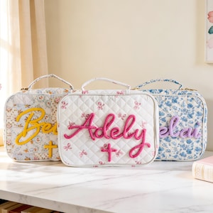 Puede incluir: Tres loncheras con estampado floral con nombres y cruces bordados. Las loncheras presentan diferentes patrones florales en blanco, azul y una mezcla de colores. Cada lonchera tiene un asa y un nombre bordado en un color contrastante.