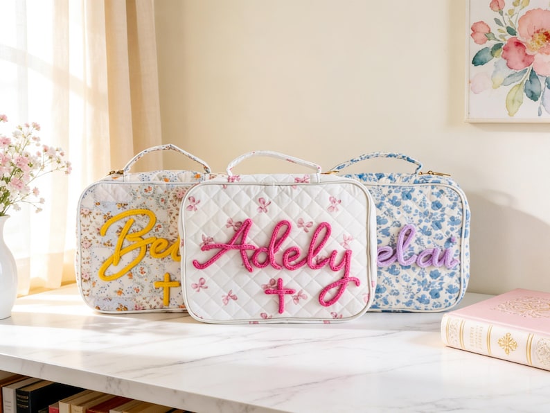 Puede incluir: Tres loncheras con estampado floral con nombres y cruces bordados. Las loncheras presentan diferentes patrones florales en blanco, azul y una mezcla de colores. Cada lonchera tiene un asa y un nombre bordado en un color contrastante.