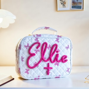 Puede incluir: Bolso de cosméticos acolchado blanco con detalles de mariposas rosas y el nombre "Ellie" bordado en rosa intenso. El bolso tiene un asa superior y cierre de cremallera. Una cruz rosa está bordada debajo del nombre.