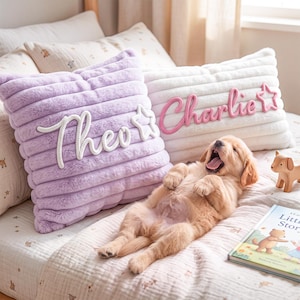 May include: Two square plush pillows with horizontal ribbed texture. One pillow is light purple with the white embroidered name Theo and a star. The other is white with the pink embroidered name Charlie and a star. A golden puppy lies in front.