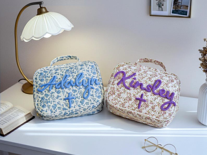 Puede incluir: Dos neceseres con estampado floral, uno azul y otro beige. La bolsa azul tiene el nombre "Adalyn" bordado en azul, y la bolsa beige tiene el nombre "Kinley" bordado en morado. Cada bolsa tiene un asa.