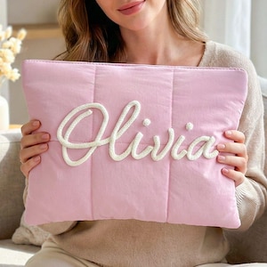 May include: A light pink quilted clutch with the name "Olivia" embroidered in white rope-like letters. The clutch has a rectangular shape and a soft, padded texture, perfect for carrying essentials.