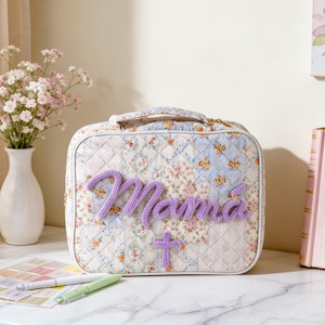 Puede incluir: Un neceser acolchado con un dise&ntilde;o de patchwork floral. La palabra "Mam&aacute;" est&aacute; bordada en hilo morado en la parte delantera, junto con una peque&ntilde;a cruz. La bolsa tiene un asa y una cremallera dorada. Un jarr&oacute;n con flores y un libro rosa est&aacute;n en el fondo.