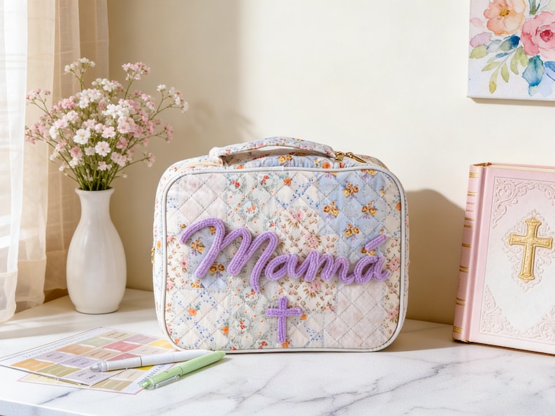 Puede incluir: Un neceser acolchado con un dise&ntilde;o de patchwork floral. La palabra "Mam&aacute;" est&aacute; bordada en hilo morado en la parte delantera, junto con una peque&ntilde;a cruz. La bolsa tiene un asa y una cremallera dorada. Un jarr&oacute;n con flores y un libro rosa est&aacute;n en el fondo.