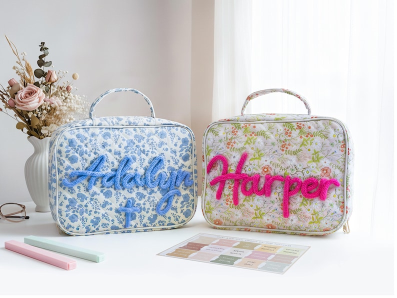 Puede incluir: Dos bolsas acolchadas personalizadas. Una es floral azul con "Adalyn" en hilo azul, la otra es floral con "Harper" en hilo rosa. Ambas tienen asas y est&aacute;n sobre una superficie blanca con muestras de color.
