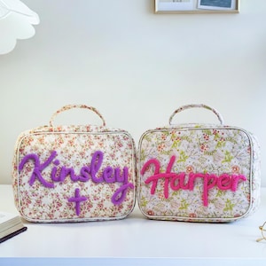 Puede incluir: Dos neceseres con estampado floral y asas. Uno tiene el nombre "Kinsley" en escritura morada, y el otro "Harper" en rosa. Ambos tienen una cruz o un signo m&aacute;s.