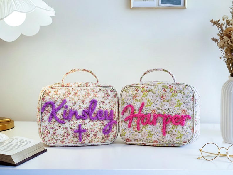 Puede incluir: Dos neceseres con estampado floral y asas. Uno tiene el nombre "Kinsley" en escritura morada, y el otro "Harper" en rosa. Ambos tienen una cruz o un signo m&aacute;s.