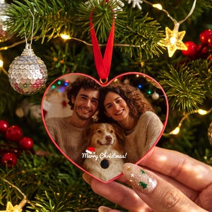 Op de afbeelding: Een hartvormige kerstversiering met een afgedrukte foto van een glimlachend koppel en een hond. Het ornament heeft een rode rand, een rood lint om op te hangen en de tekst Merry Christmas met een klein icoontje van een kerstmuts erop.