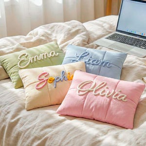 Könnte beinhalten: Fünf rechteckige Kissen in Pastellfarben, jedes mit einem Namen in Schreibschrift bestickt. Die Kissen sind grün, hellblau, pfirsichfarben und rosa. Sie liegen auf einem Bett mit einem Laptop und einer Tasse Kaffee.