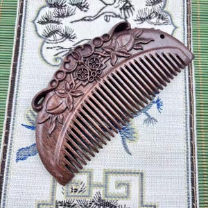 May include: A brown wooden comb with detailed floral and geometric carvings. The comb has fine teeth and a decorative handle with relief patterns. The comb is displayed on patterned fabric.