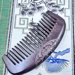 Op de afbeelding: Een donkerbruine houten kam met dicht op elkaar geplaatste tanden. De kam heeft een gebogen handvat met een geëtst decoratief ontwerp. De kam ligt op een stof met een patroon met een groene en witte achtergrond.