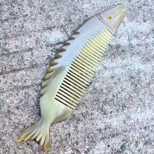 May include: A fish-shaped comb with a light brown, translucent body. The comb features a detailed fish design, including fins, scales, and an eye. The comb's teeth are a lighter shade of yellow. The comb is made of a smooth material.