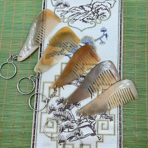 May include: Five horn combs with keyrings. The combs are in shades of cream, brown, and tan. They are displayed on a decorative cloth, set against a green bamboo mat.