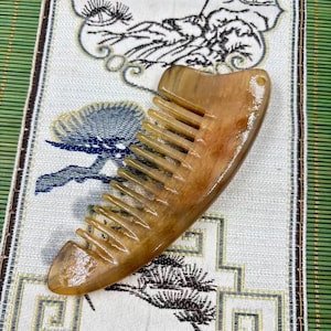 May include: A light brown, horn comb with a curved handle and evenly spaced teeth. The comb has a smooth, polished surface and a small hole at the top. The comb is laying on a patterned fabric.