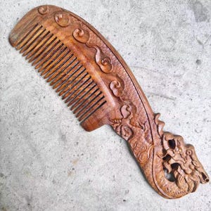 May include: A wooden comb with intricate dragon carvings. The comb is made of a dark brown wood and features fine teeth. The handle is carved with a detailed dragon design, showcasing scales and a dragon head. The comb is approximately 7 inches long.