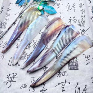 May include: Five handcrafted combs with a horn-like appearance. The combs have a gradient of colors, including brown, purple, and cream. The teeth are finely spaced. The combs are arranged on a patterned fabric.