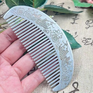 May include: A silver-colored comb with a curved design. The comb features detailed carvings of fish and cloud patterns. The teeth of the comb are evenly spaced. The comb is held in a hand.
