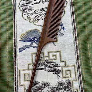 May include: A dark brown wooden hair comb featuring a long, pointed tail handle and fine teeth. The comb has a smooth, polished finish and a slender, tapered design, showcasing the natural grain of the wood throughout its entire structure.