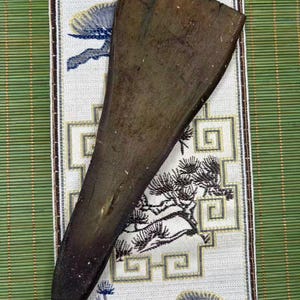 May include: A dark brown, wedge-shaped wooden tool with a smooth, flat surface and a tapered end. The object features a natural wood grain texture with visible variations in colour, ranging from deep espresso to lighter brown tones along the edges.