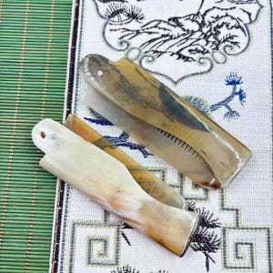 May include: Two horn-handled straight razors. One razor is open, revealing the blade and teeth. The other razor is closed. The handles are a mix of light and dark brown, with a glossy finish. The razors rest on a patterned fabric.