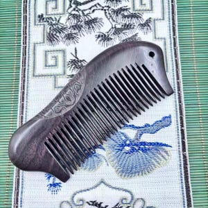 May include: A dark brown wooden comb with fine teeth. The comb features an engraved floral design on the handle and a small hole near the top. The comb is displayed on a patterned cloth with blue and white floral motifs.