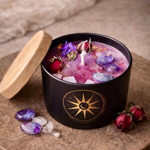 Könnte beinhalten: Eine schwarze Kerze in einem runden Behälter mit Holzdeckel, verziert mit einem goldenen Sonnen- und Mondemblem. Das violette Wachs enthält eingebettete violette und klare Kristalle, getrocknete Rosenknospen und blaue Blütenblätter mit einem Docht.
