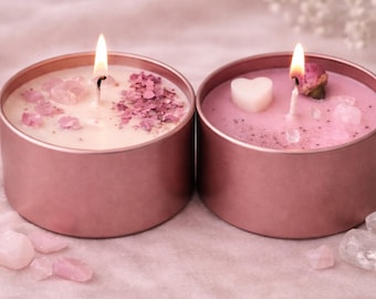 May include: Two round rose gold metal tins containing candles with lit wicks. The left candle is white with pink crystal fragments and petals. The right candle is pink with a white heart-shaped wax piece, a dried rosebud, and clear crystals.