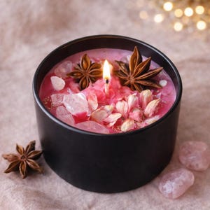 Könnte beinhalten: Eine brennende, rosa Kerze in einem schwarzen Behälter. Die Kerze ist mit Sternanis, Rosenquarzkristallen und getrockneten Blumen verziert. Die Kerze steht auf einer neutralfarbenen Oberfläche.