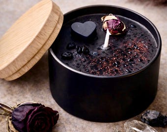 May include: A black candle in a round metal tin with a light wood lid. The black wax surface is decorated with a small black heart, a dried rosebud, copper-colored glitter, and small black stones. A single white wick stands in the center.