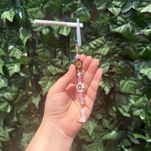 May include: A metal roach clip featuring a beaded strand with pink, clear, and brown floral-shaped beads, finished with a white cowboy boot charm. The clip holds a rolled cigarette, showcasing a decorative design with various bead textures and colours.