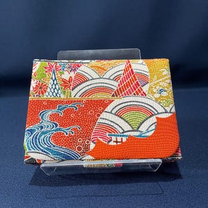 以下が含まれることがあります： 日本のデザインにインスパイアされたカラフルな財布。財布は、赤、青、オレンジ、黄色、緑色の波、山、花の要素の鮮やかなパターンが特徴です。財布は、濃い青色の背景に対して透明なプラスチックスタンドに表示されています。