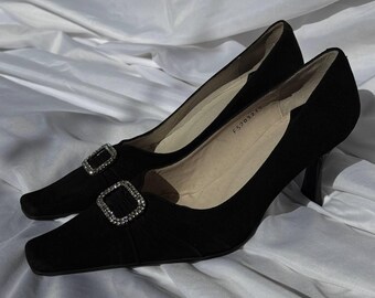 Könnte beinhalten: Ein Paar schwarze Wildleder-Pumps mit quadratischen Zehen und kleinen Absätzen. Jeder Schuh hat eine dekorative quadratische Schnalle mit funkelnden Verzierungen. Die Schuhe stehen vor einem weißen, drapierten Stoffhintergrund.