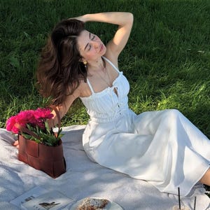 Puede incluir: Una escena de picnic con un atuendo blanco, una bolsa de cuero trenzado marr&oacute;n llena de flores rosas y un plato de cruasanes. La escena est&aacute; ambientada en una manta a rayas grises y blancas sobre un c&eacute;sped. Tambi&eacute;n hay dos bebidas heladas y un libro.