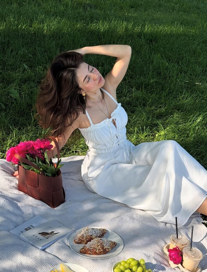 Puede incluir: Una escena de picnic con un atuendo blanco, una bolsa de cuero trenzado marr&oacute;n llena de flores rosas y un plato de cruasanes. La escena est&aacute; ambientada en una manta a rayas grises y blancas sobre un c&eacute;sped. Tambi&eacute;n hay dos bebidas heladas y un libro.