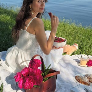 Puede incluir: Una escena de picnic con una manta blanca sobre hierba cerca del agua. Una bolsa tote de cuero trenzado marr&oacute;n contiene peon&iacute;as de color rosa brillante. Un cuenco blanco con fresas, cruasanes y una bebida tambi&eacute;n est&aacute;n sobre la manta.