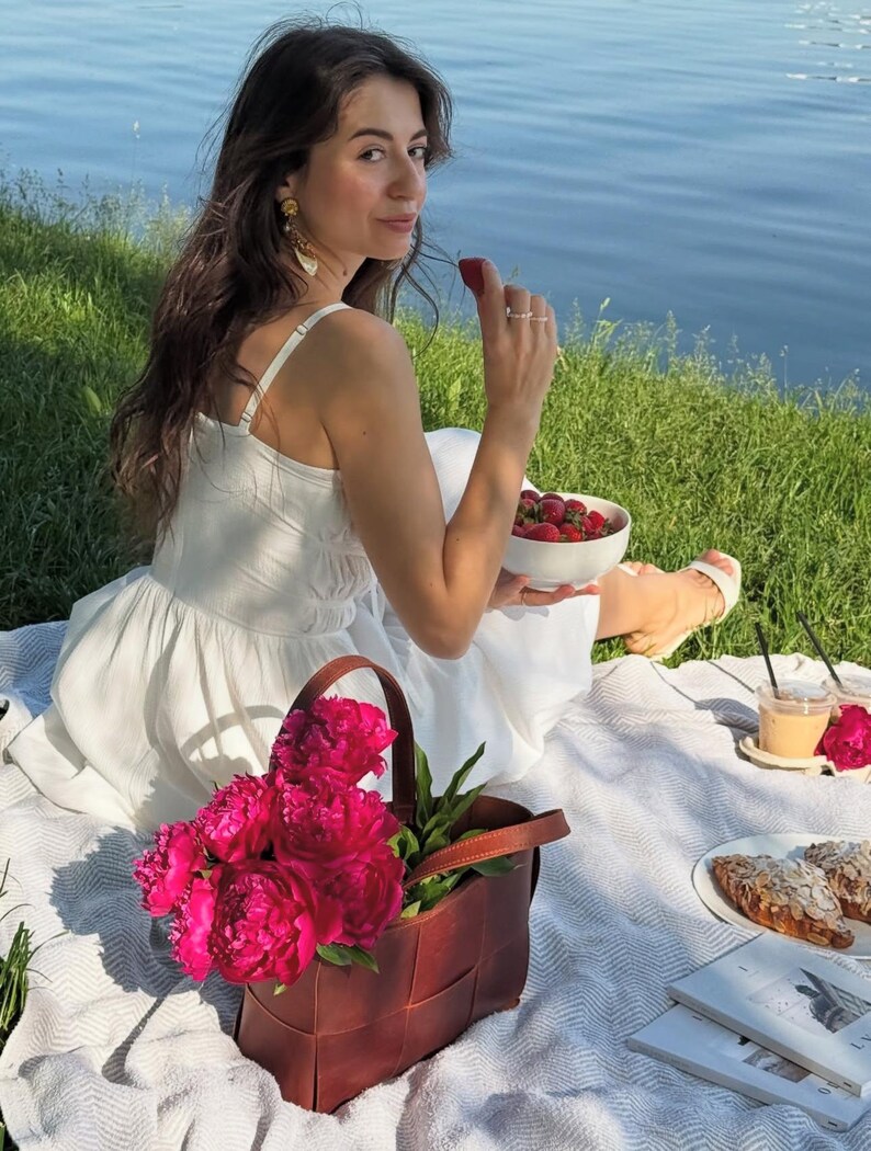 Puede incluir: Una escena de picnic con una manta blanca sobre hierba cerca del agua. Una bolsa tote de cuero trenzado marr&oacute;n contiene peon&iacute;as de color rosa brillante. Un cuenco blanco con fresas, cruasanes y una bebida tambi&eacute;n est&aacute;n sobre la manta.