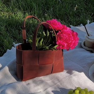 Puede incluir: Un bolso tote de cuero marr&oacute;n con dise&ntilde;o tejido, lleno de vibrantes peon&iacute;as rosas. El bolso tiene dos asas y est&aacute; sobre una manta estampada blanca y gris, con un cuenco de uvas verdes y una bebida cerca.