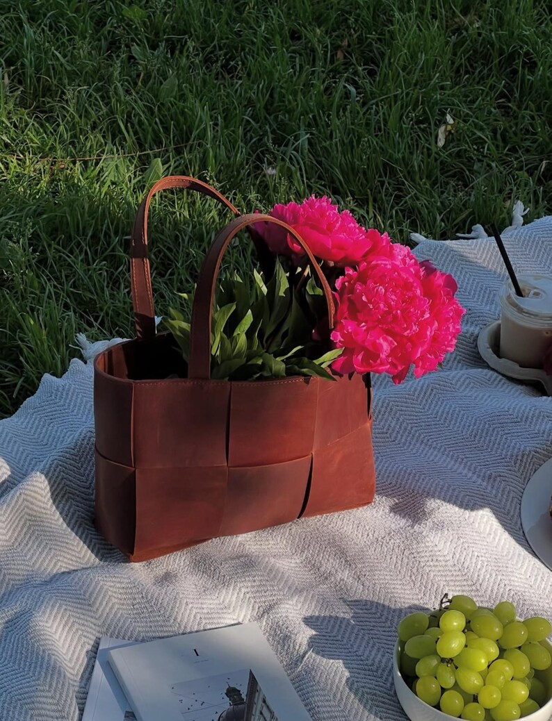 Puede incluir: Un bolso tote de cuero marr&oacute;n con dise&ntilde;o tejido, lleno de vibrantes peon&iacute;as rosas. El bolso tiene dos asas y est&aacute; sobre una manta estampada blanca y gris, con un cuenco de uvas verdes y una bebida cerca.