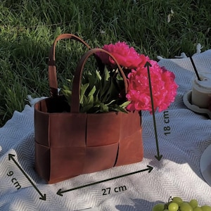 Puede incluir: Una cesta de cuero marr&oacute;n con dise&ntilde;o tejido, llena de vibrantes flores rosas. La cesta, de 27 cm de largo, 9 cm de ancho y 18 cm de alto, tiene dos asas. La escena est&aacute; ambientada al aire libre sobre una manta con uvas y una bebida.