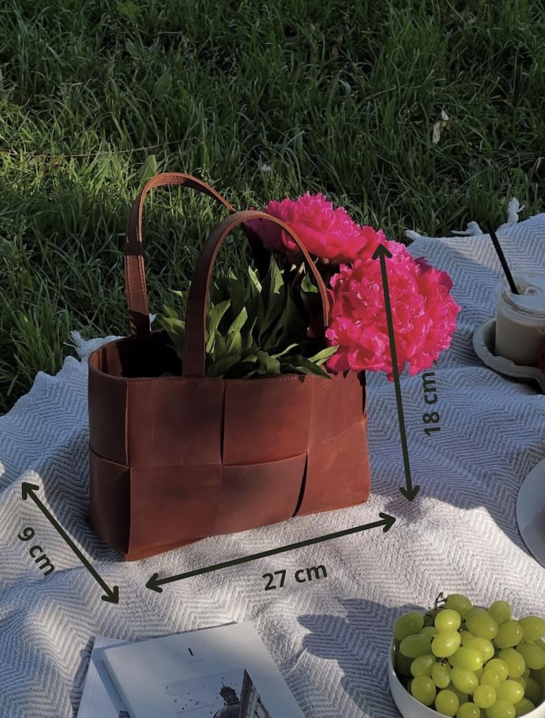 Puede incluir: Una cesta de cuero marr&oacute;n con dise&ntilde;o tejido, llena de vibrantes flores rosas. La cesta, de 27 cm de largo, 9 cm de ancho y 18 cm de alto, tiene dos asas. La escena est&aacute; ambientada al aire libre sobre una manta con uvas y una bebida.