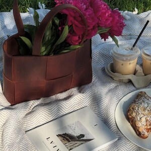 Puede incluir: Escena de picnic con un bolso tote de cuero marr&oacute;n lleno de peon&iacute;as rosas. Dos caf&eacute;s helados con pajitas, un cruas&aacute;n y un libro con la palabra "LVIV" sobre una manta blanca.
